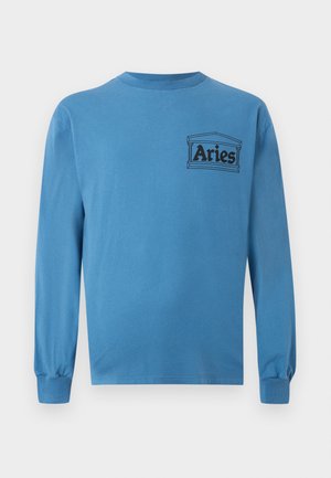Homme portant des lunettes de soleil noires, une chemise bleue à manches longues avec le logo "Aries", un short large noir, des bottes beige à lacets, mains dans les poches, debout sur fond blanc.