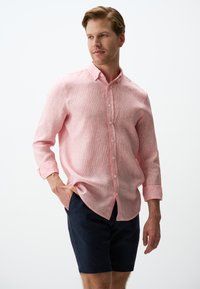 Helles rosa Leinenhemd mit kleinen weißen Streifen, lange Ärmel, die an den Manschetten hochgerollt sind, getragen mit dunkelblauen Shorts.