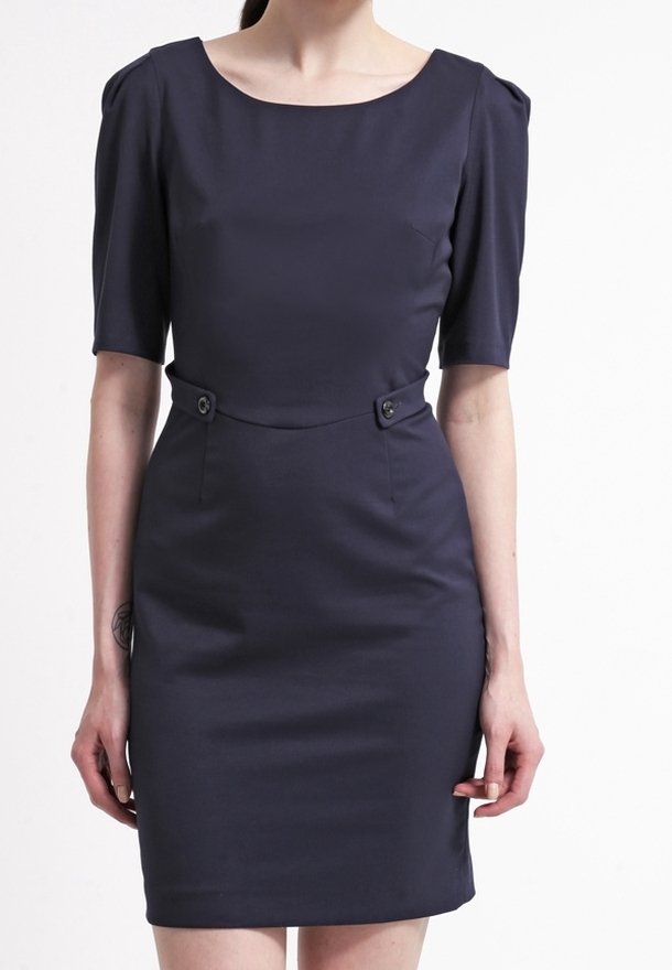 Femme portant une robe bleu marine ajustée avec des manches au coude et des détails de boutons à la taille, debout devant un fond uni.