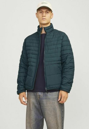 Pudret teal jakke med høj krave, lynlåslukning og quiltet tekstur. Bæres over en mørk sweater og kombineres med lys denim.