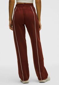 Sarkani sporta bikses ar brīvu piegriezumu, kurām ir baltas vertikālas svītras un elastīga josta. Gluda tekstūra, piemērota sporta apģērbam.