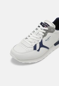 Weißes Leder-Sneaker mit marineblauen und grauen Akzenten, durchlochter Zehenpartie und weißen Schnürsenkeln. Es hat ein Logo an der Seite und eine gepolsterte Sohle.