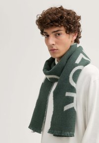 Ciemnozielony wełniany szalik z kontrastowym jasnoszarym logo. Frędzlowe krawędzie i miękka tekstura dodają detalu. Noszony luźno na szyi.