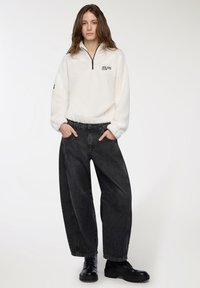 Giovane vestito con un fleece bianco a zip quarti con il logo "New York Athletics Club", jeans neri larghi e stivali neri con lacci, in piedi con le mani in tasca.