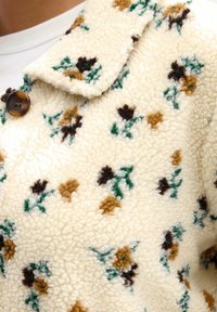 Kreemivärvi fleece-jakk, millel on pruunide, mustade ja roheliste lillede mustriga kaunistus, kaelus ja ümmargune pruun nööbikas.