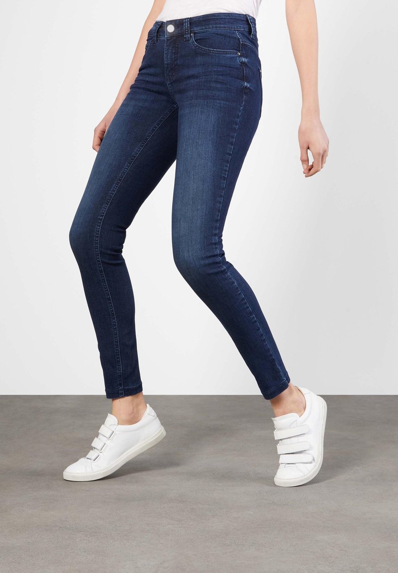 Mörkblå skinny jeans i denim med medelhög midja, femfickorsdesign och synlig sömnad. Bärs med vita sneakers.