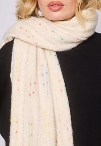 Écharpe crème en tissu doux, ornée de taches colorées dans différentes teintes, enroulée autour du cou, avec une texture côtelée.