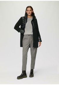 Manteau noir avec un panneau avant matelassé gonflé, pull en tricot gris, pantalon gris et bottines noires avec des accents de semelle verte. Design simple, texture élégante.