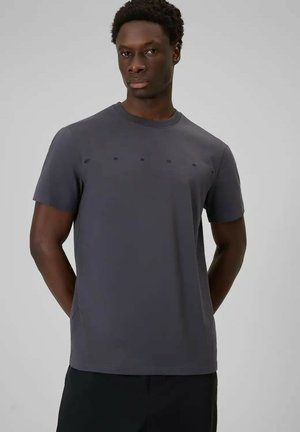 Mężczyzna o ciemnej karnacji, ubranego w krótki szary t-shirt koloru węgla drzewnego z subtelnym, tonowym napisem, stojącego na jednolitym jasnoszarym tle.