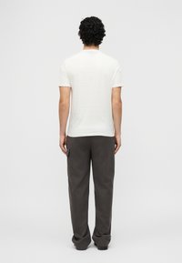 Άνδρας με σγουρά μαλλιά στέκεται με την πλάτη γυρισμένη, φοράει απλό λευκό κοντομάνικο μπλουζάκι και σκούρα γκρι φαρδιά παντελόνια με πλαϊνή τσέπη.