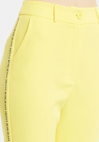 Pantaloni gialli realizzati in tessuto liscio, con tasche laterali e chiusura a bottone. Un logo nero a contrasto corre verticalmente lungo il lato.