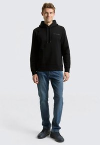 Black hooded sweatshirt with drawstrings, front kangaroo pocket, and logo detail. Worn with blue jeans and dark lace-up shoes.