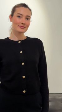 Jauna moteris su surištais plaukais, dėvinti juodą viršutinį drabužį su aukso spalvos širdies formos sagomis, stovinti priešais paprastą šviesų foną.