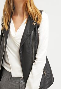 Schwarze Lederweste mit lockerem Schnitt, silbernen Reißverschlüssen, kombiniert mit einem weißen Rollkragen-Top und grauen Hosen.