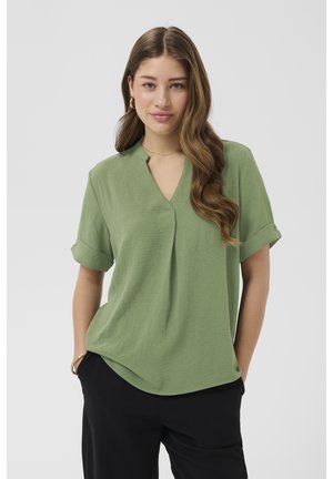 Jeune femme aux longs cheveux ondulés portant un chemisier vert à manches courtes et un pantalon noir, debout les mains dans les poches sur un fond uni.