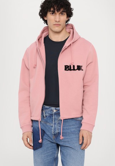 Rozā zip-up kapuce ar auklām, piedāvā teksturētu "BLUE" uzrakstu un rotājumus. Uzvilkta virs tumša krekliem un apvienota ar džinsiem.