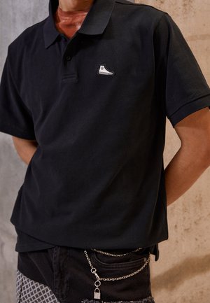 Czarna koszulka polo z krótkim rękawem z małym białym logo sneakersa na piersi, noszona z czarnymi spodniami i srebrnym łańcuszkiem z kłódką.