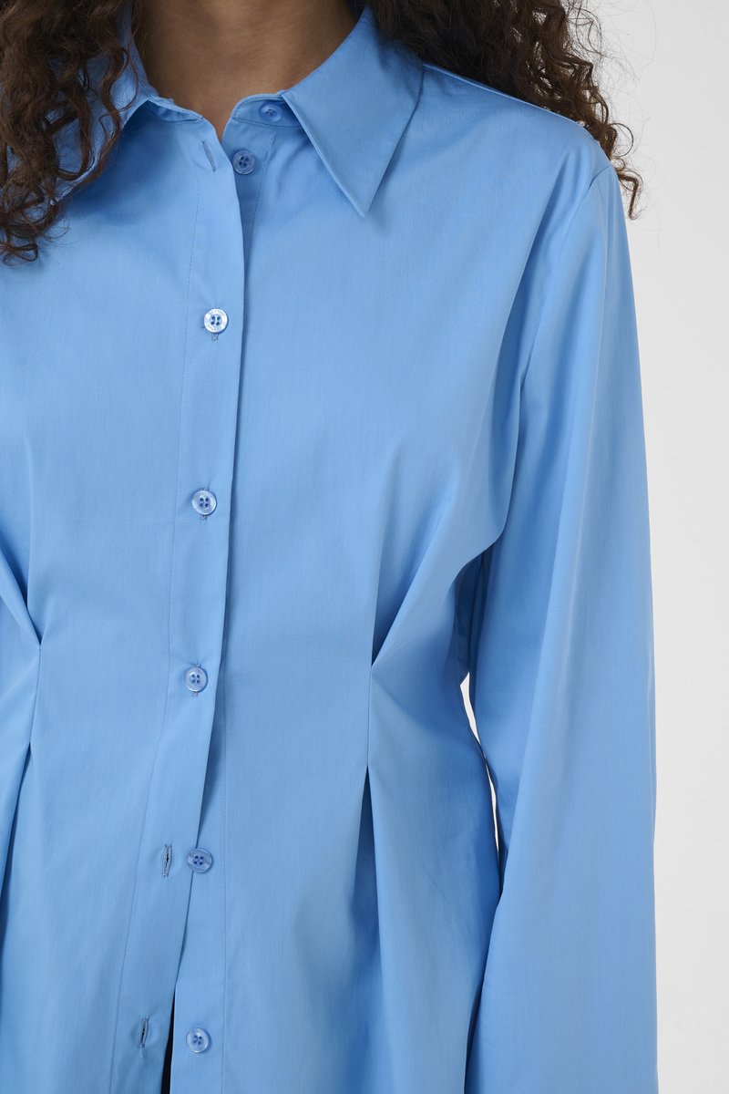 Femme portant une chemise cintrée bleu clair à boutons, manches longues et col large, vue du cou à la taille.