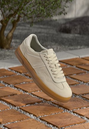 Beżowy sneaker Tommy Jeans z gumową podeszwą i sznurowadłami na drewnianych kwadratowych kafelkach z białymi kamykami i rozmytym drzewem w tle.