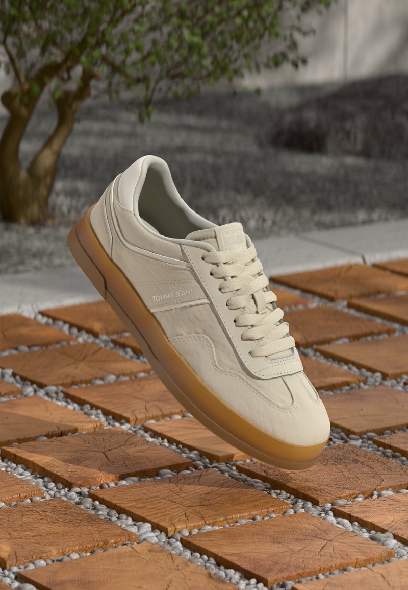 Baskets beige Tommy Jeans avec semelle en gomme et lacets sur des carreaux de bois carrés avec des cailloux blancs et un arbre flou en arrière-plan.