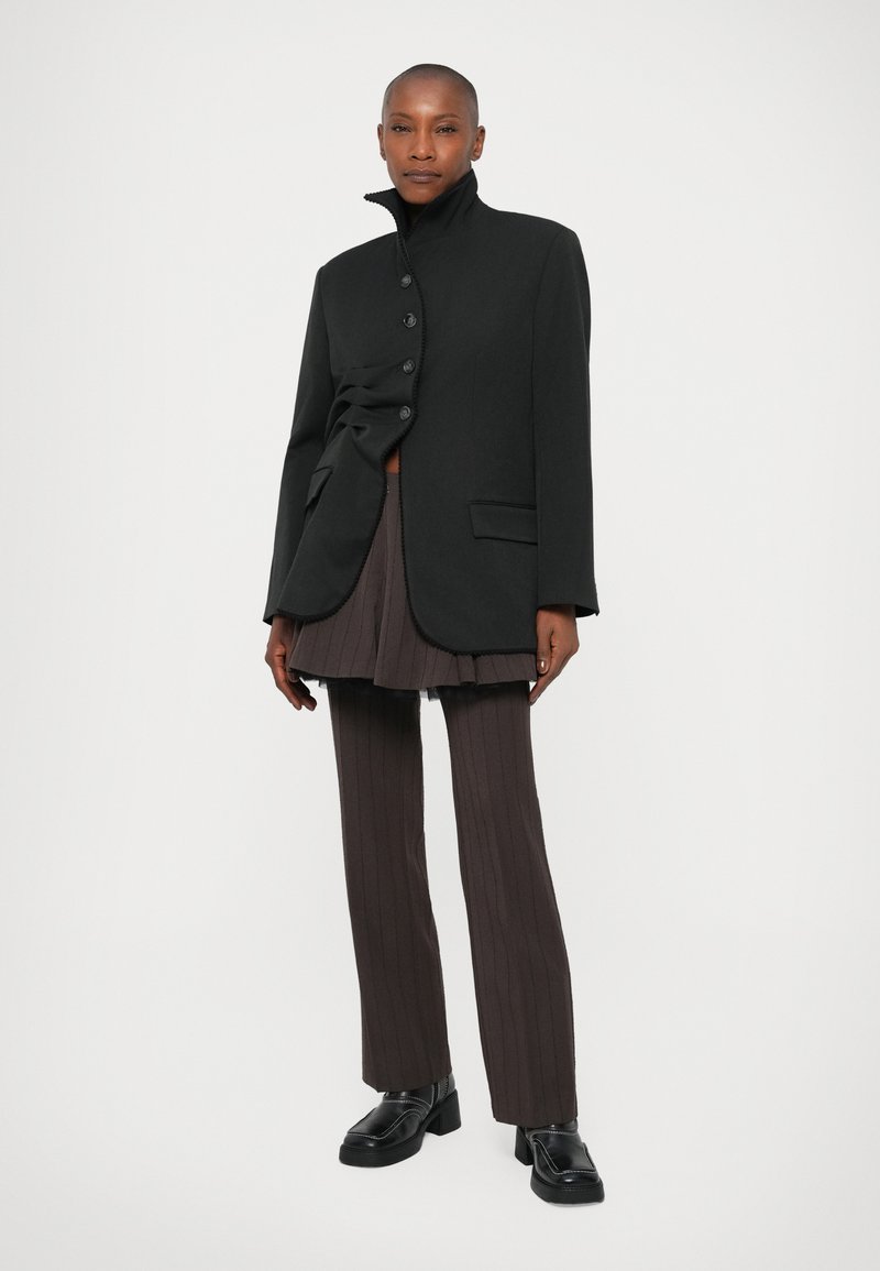 Schwarze taillierte Jacke mit hohem Kragen und vier Knöpfen, kombiniert mit dunkelgrauen Pliselhosen. Klare Linien und strukturiertes Design.