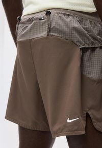 Κοντινό πλάνο σε καφέ σορτς Nike με καρό μέση, τσέπη με φερμουάρ, κορδόνι και λευκό λογότυπο Nike κοντά στο τελείωμα.