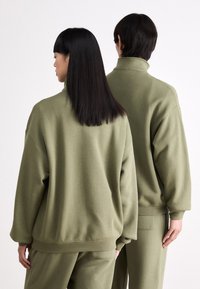 Sudaderas oversized color verde oliva con cuellos altos que presentan un tejido suave, hombros caídos y puños elásticos, llevadas por dos figuras vistas desde atrás.