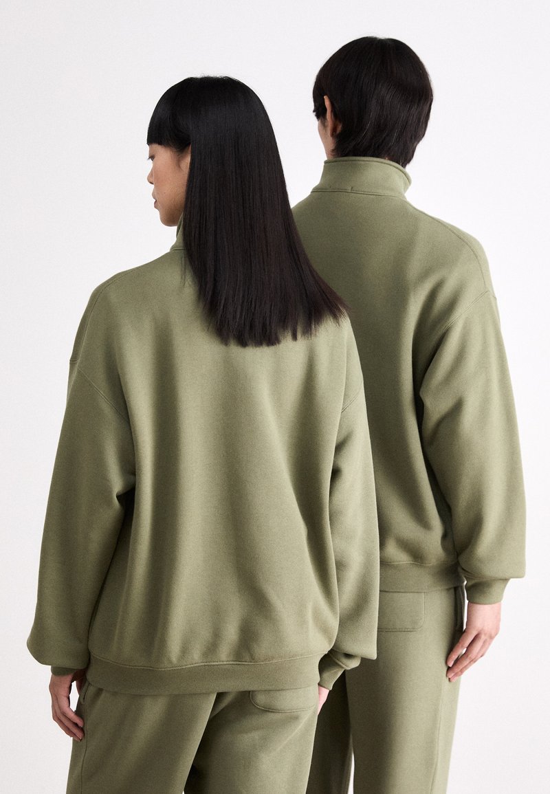 Des sweatshirts surdimensionnés vert olive avec des cols hauts présentent un tissu doux, des épaules tombantes et des poignets élastiques, portés par deux silhouettes vues de dos.
