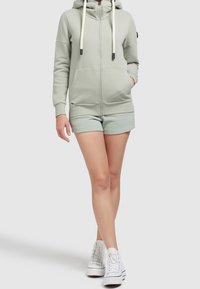 Persona che indossa una felpa verde chiaro con zip e tasche, pantaloni corti abbinati e sneakers bianche alte, in piedi contro uno sfondo semplice.