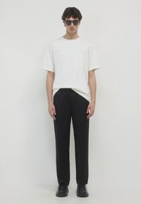 T-shirt bianco a maniche corte con taschino, abbinato a pantaloni neri su misura. Il modello indossa occhiali da sole neri e scarpe scure. Design semplice, colori solidi.