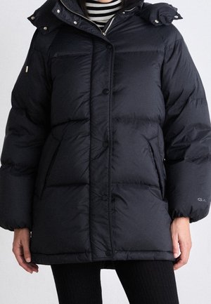 Nainen, joka pitää yllään mustaa tikattua toppatakkia, jossa on painonapit, sivutaskut ja huppu, yhdistettynä mustiin resorihousuihin ja raidalliseen toppiin.