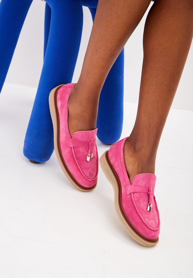 Mocassins en suede rose avec une texture douce, dotés d'un accent métalleux décoratif et d'une semelle en caoutchouc brun clair, posés sur une chaise bleue.