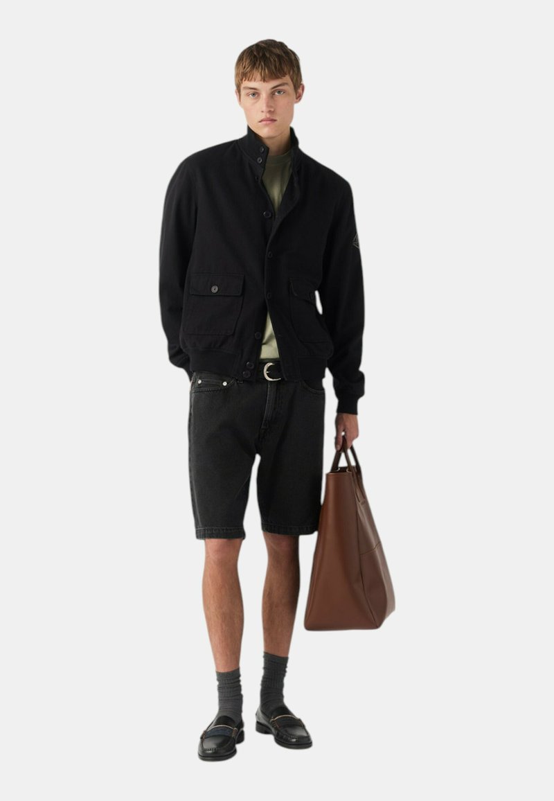 Giacca nera con bottoni, colletto alto, t-shirt verde, pantaloni shorts in denim neri, borsa a tracolla in pelle marrone e scarpe nere con calze.