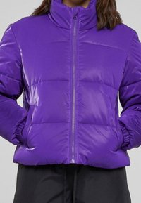 Fioletowa kurtka puchowa z wysokim kołnierzem, zamkiem błyskawicznym z przodu, elastycznymi mankietami i pikowaną teksturą. Materiał wydaje się błyszczący i ocieplający.
