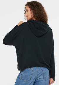 Schwarzer, strukturierter Kapuzenpullover mit lockerer Passform, geripptem Muster und lässigem Design, kombiniert mit hellblauen, hochgeschnittenen Jeans.