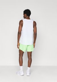 Άνδρας που στέκεται με πλάτη, φορώντας ένα λευκό αμάνικο πουκάμισο, ανοιχτού πράσινου αθλητικά σορτσάκια, λευκές κάλτσες και λευκά αθλητικά παπούτσια, σε ένα απλό φόντο.