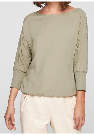 Vaaleanoliivinvärinen väljälahkeinen pitkähihaisen pusero kalvosin ja koristepainikkein ylähihoissa, yhdistetty vaaleanbeigeihin housuihin.