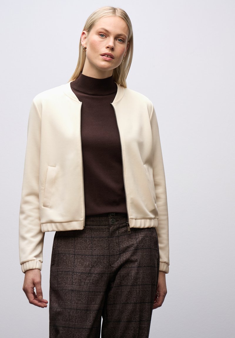 Giacca bomber zip-up crema con polsini a coste, indossata sopra un dolcevita marrone scuro. Pantaloni a vita alta a scacchi scuri con due bottoni.