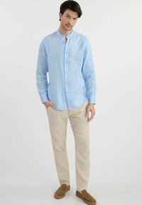 Jasnoniebieska koszula z długim rękawem na guziki z kołnierzem mandarynkowym, w zestawie z beżowymi spodniami i brązowymi butami wsuwanymi.