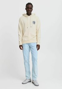Sweat à capuche crème avec un graphique sur le devant, associé à un jean bleu clair et des baskets grises, offrant une coupe décontractée et une poche kangourou.