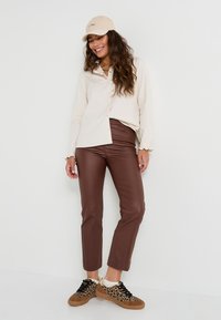 Camisa beige de botones, pantalones de imitación cuero marrón y zapatillas con estampado de leopardo. La gorra es de color marrón claro con bordado. Combinación de atuendo casual.