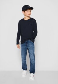 Camicia a maniche lunghe blu navy, jeans di denim, sneakers bianche. Un cappello scuro con il logo "TOMMY HILFIGER" completa l'outfit. Design semplice e casual.