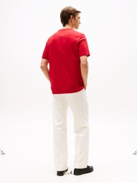 Rotes Baumwoll-T-Shirt mit lockerer Passform, kurzen Ärmeln und rundem Ausschnitt, kombiniert mit weißen Hosen und schwarzen Schuhen, von hinten gezeigt.