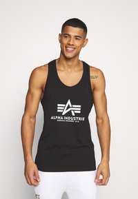 Schwarzes Tanktop mit einem großen weißen Logo, das "ALPHA INDUSTRIE" mit einem geometrischen Design zeigt. Der Stoff wirkt glatt und enganliegend.