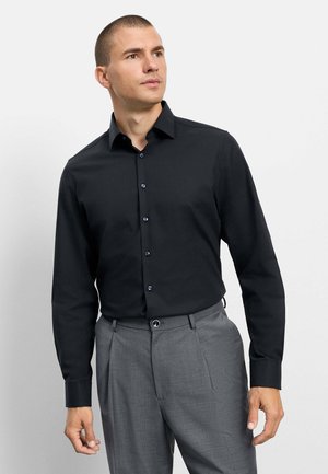 Man wearing a black long-sleeve button-up shirt and gray dress pants, looking slightly upward and to the side.
