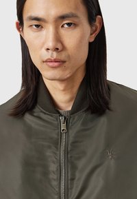 Un jeune avec de longs cheveux noirs porte une veste bombardier verte avec une fermeture éclair et un petit logo d'animal brodé sur la poitrine, sur un fond neutre.