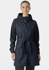 Vrouw met een lichte huid en sproeten, gekleed in een marineblauwe regenjas met capuchon, knoopsluitingen en een ceintuur, staand tegen een effen achtergrond.