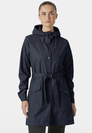 Kobieta o jasnej karnacji i piegach, ubrana w granatowy płaszcz przeciwdeszczowy z kapturem, zapinany na guziki i zawiązany paskiem, stojąca na jednolitym tle.