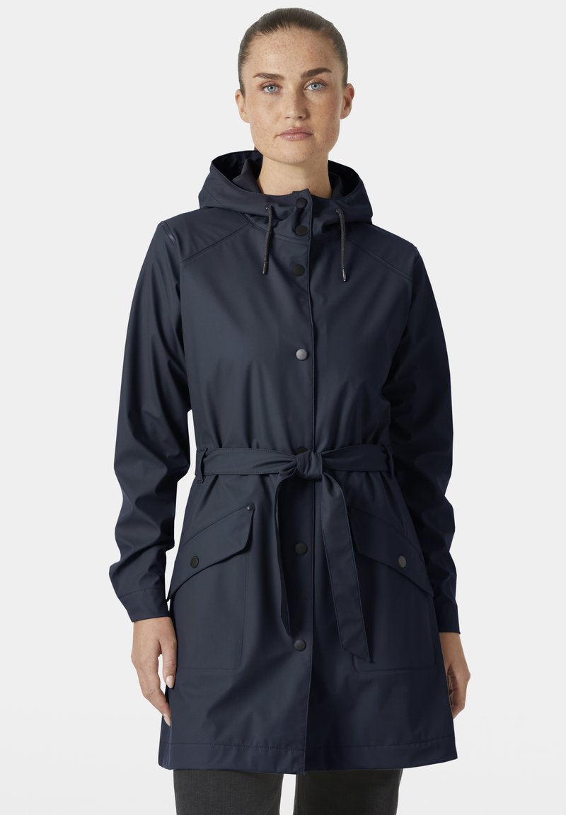 Vrouw met een lichte huid en sproeten, gekleed in een marineblauwe regenjas met capuchon, knoopsluitingen en een ceintuur, staand tegen een effen achtergrond.