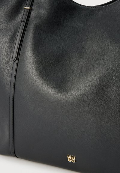 Sac à main en cuir noir avec petit logo doré "HUGO" et détail de sangle réglable sur une surface à texture lisse.